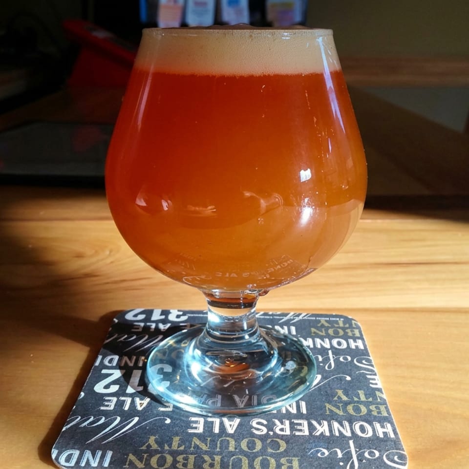Amber beer in a tulip glass sits on a typographic coaster atop a sunlit wooden restaurant table.