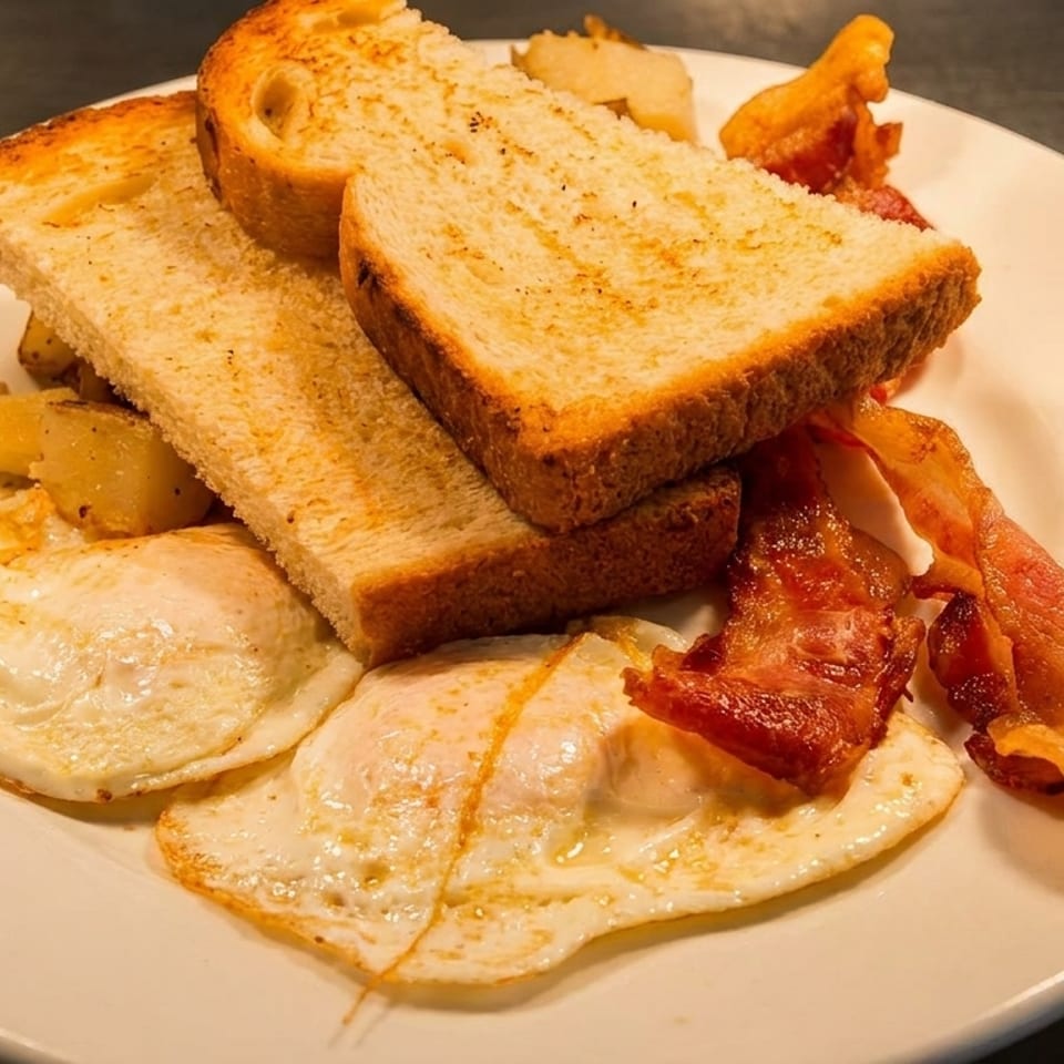 Plate of fried eggs, bacon, toast, and roasted potatoes served on a white dish.