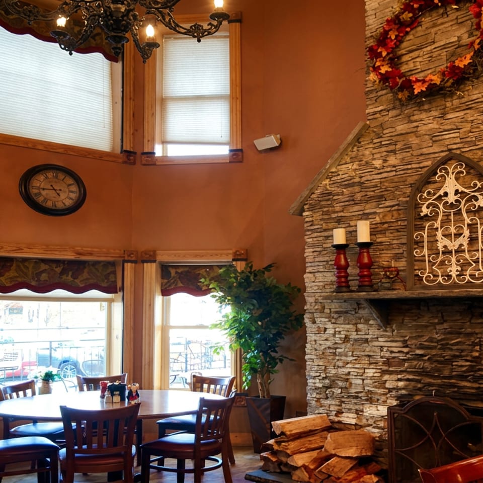 Rustic restaurant dining room with a large stone fireplace, candlelit mantel, round table, wooden chairs, and tall windows.