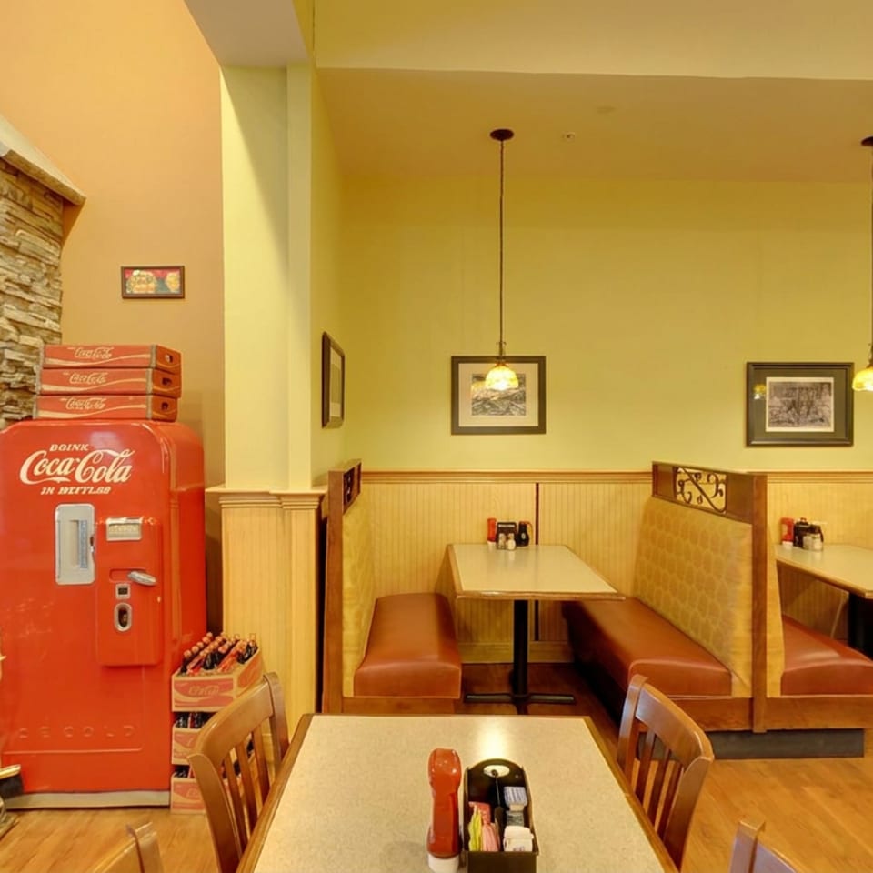 Warmly lit restaurant dining area with leather booths, wooden tables, pendant lights, stone fireplace, and vintage Coca-Cola cooler.