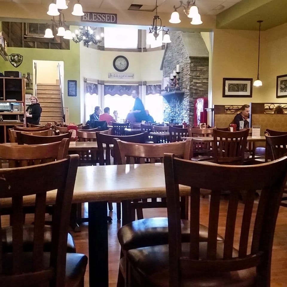 Warmly lit restaurant dining room with wooden tables, chairs, framed artwork, stone fireplace, and several diners.