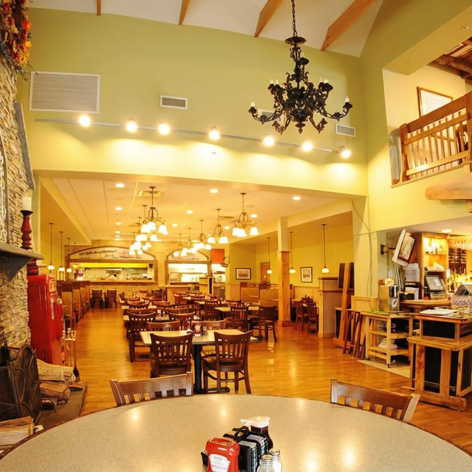 Warmly lit restaurant interior featuring a stone fireplace, wooden tables, chandeliers, a bar, and an upper-level balcony.