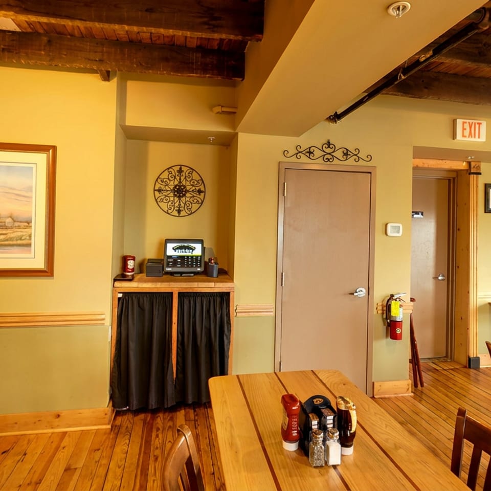 Warmly lit restaurant dining room with wooden tables, chairs, exposed beams, framed artwork, and a condiment set.