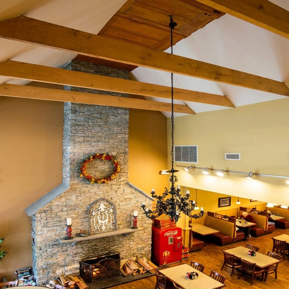 Rustic restaurant dining room with exposed timber beams, stone fireplace, chandelier, wooden tables, and booth seating.